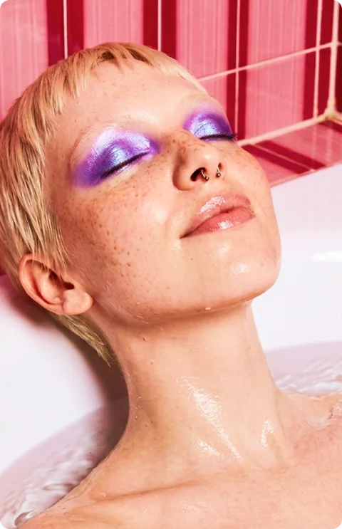 woman with purple eyeshadow relaxing, eyes closed, in a pink-tiled bath